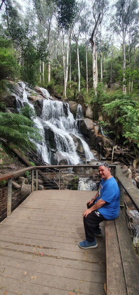 man sitting on wooden bench  on raised platform next to a beautiful waterfall with gum trees in the background